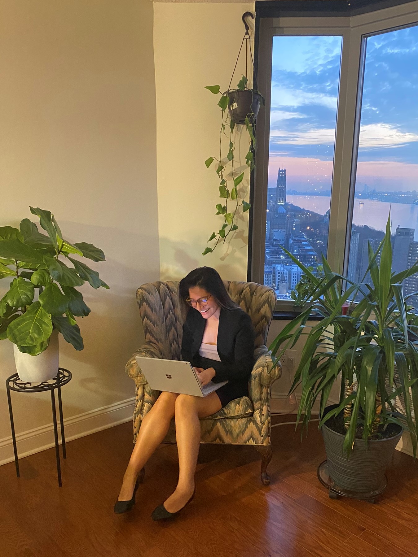 Tatiana Vera sits in an armchair on her laptop in front of a window, through which you can see the sunset over New York City