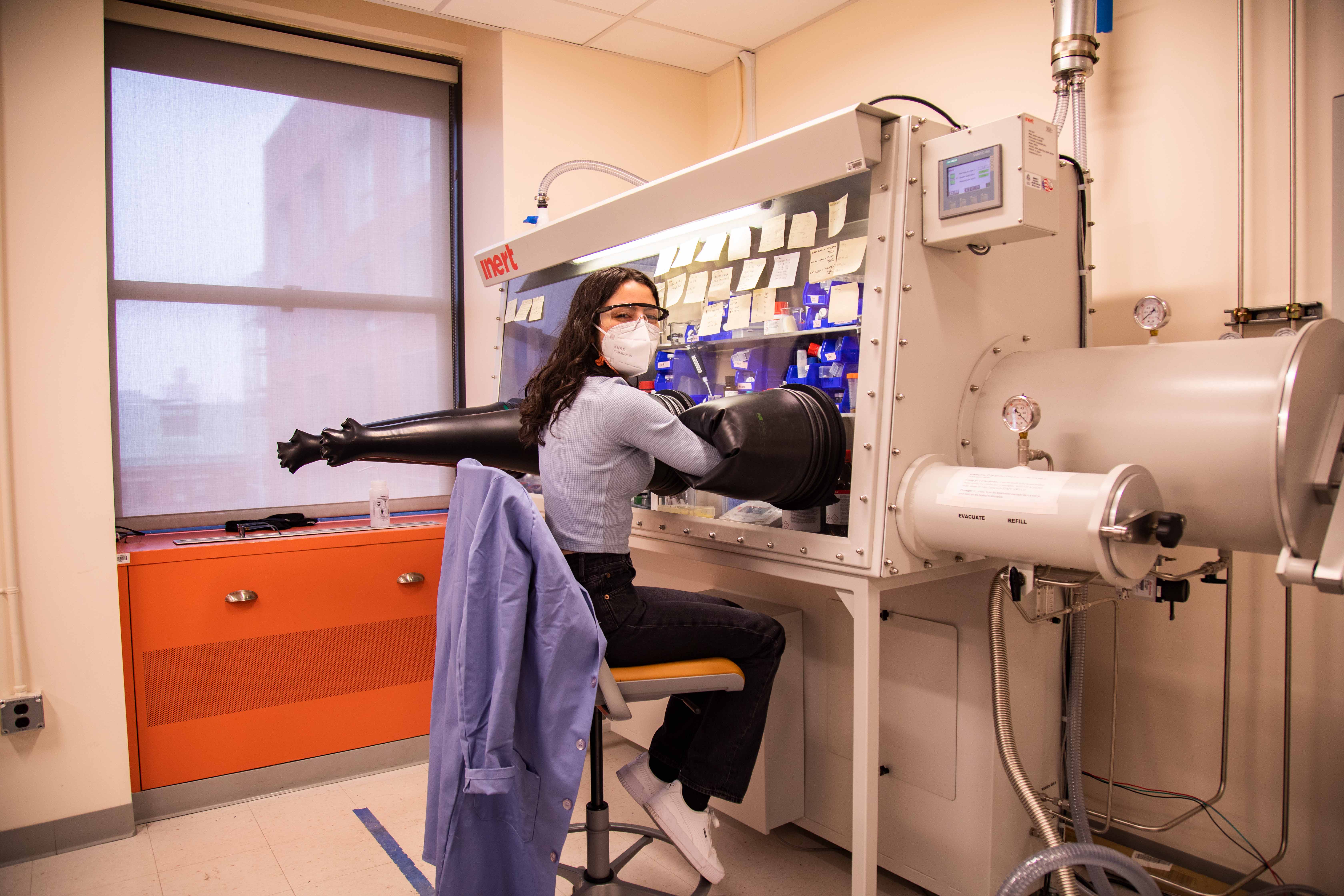 Wearing safety goggles and a protective mask, Naiara Munich looks back at the camera while her arms are inside a piece of lab equipment designed to create an air-free environment