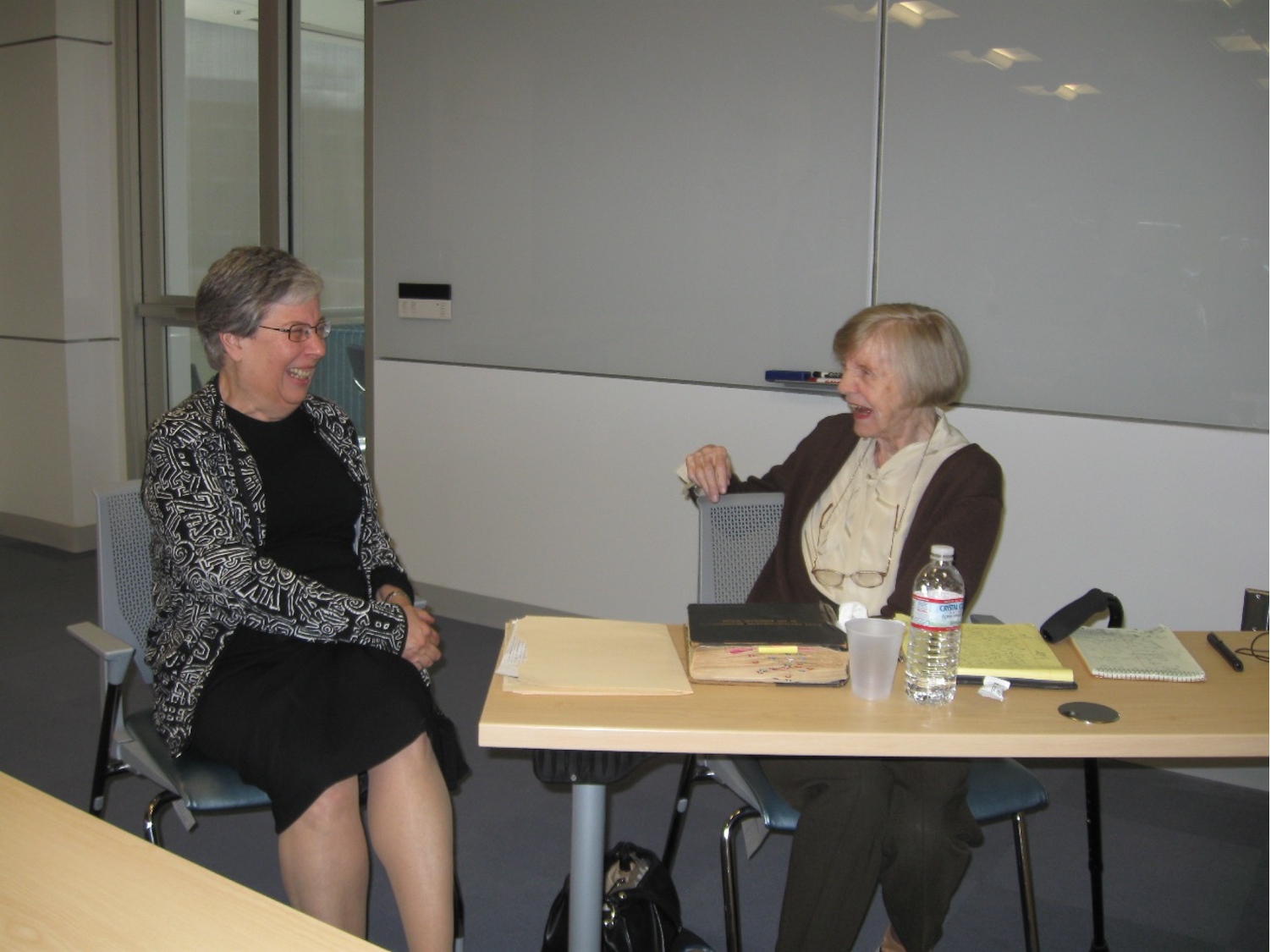 Two women sit at a desk in a classroom smiling and laughing