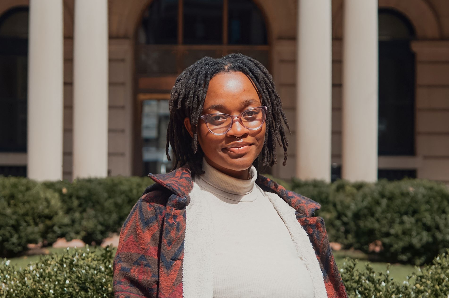 Tapiwa pictured in front of Milbank Hall wearing glasses, a white shirt, and a patterned sweater.