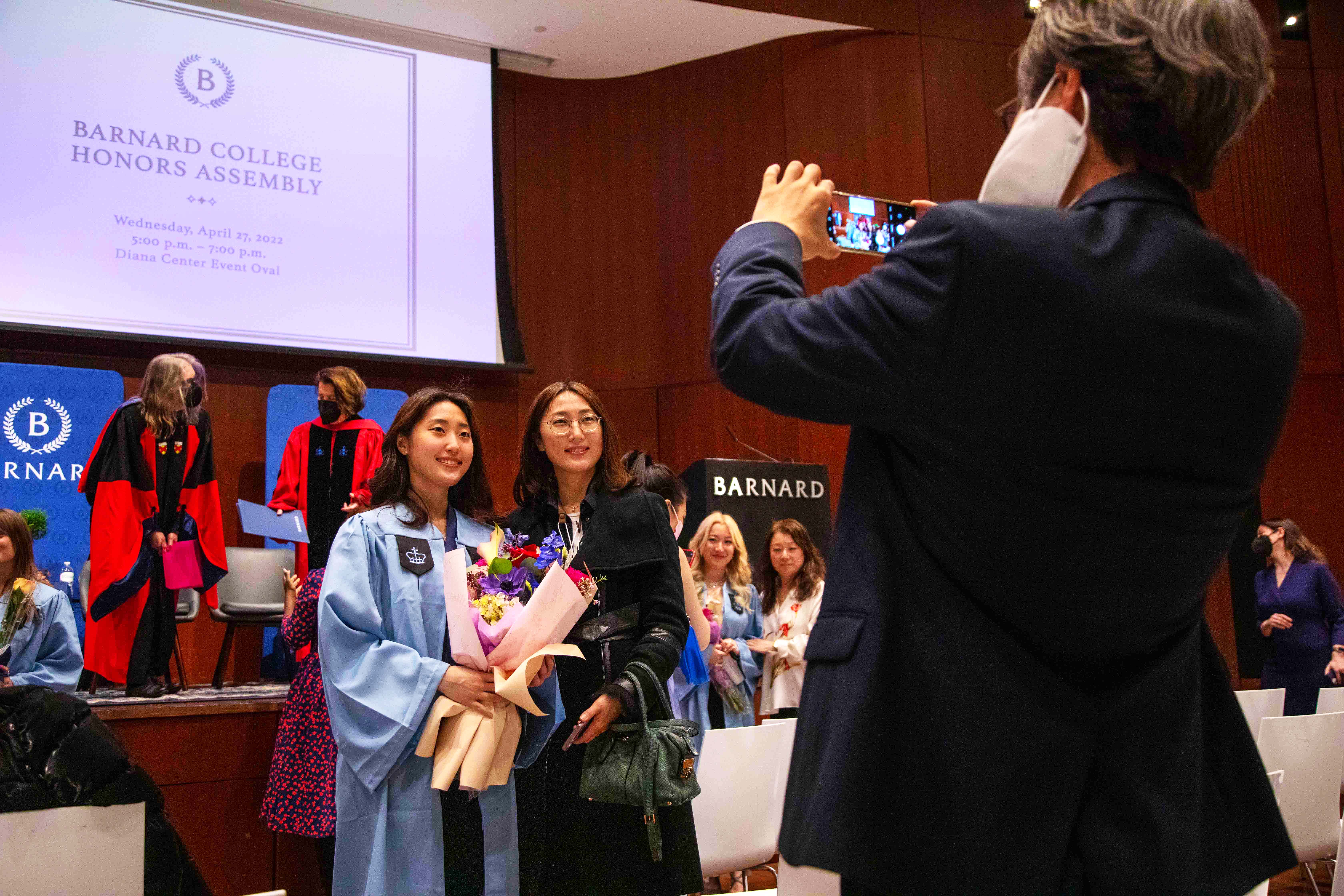 A man in a black suit jacket takes a picture of a Barnard student and her guest at the honors assembly.