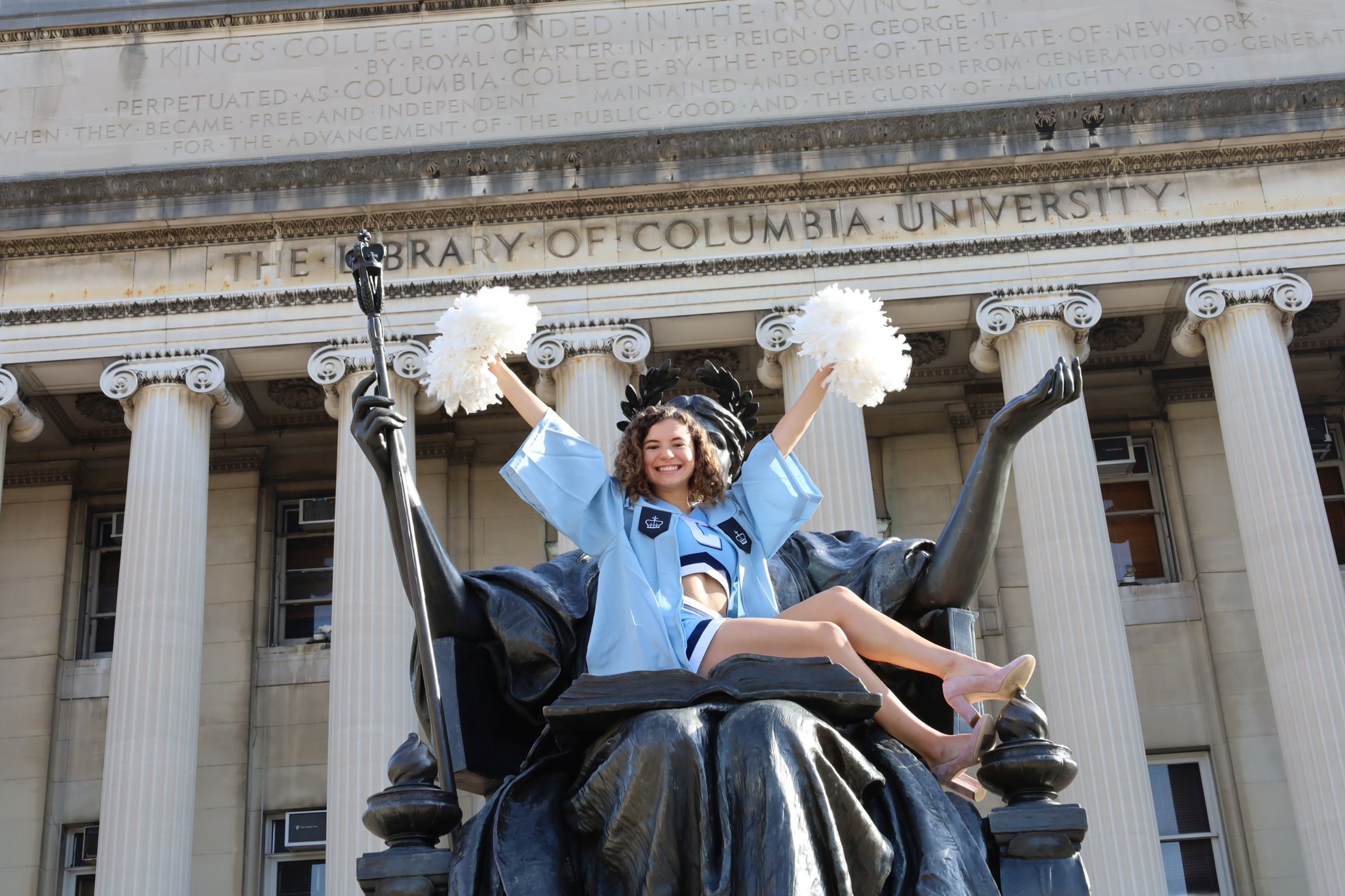 Madeleine Morales sitting on to of the Alma Mater statue in her cap and gown, holding up two white pom poms