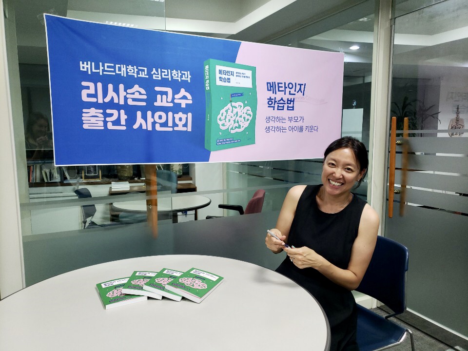 Woman wearing black dress seated at round table with banner of book behind her.