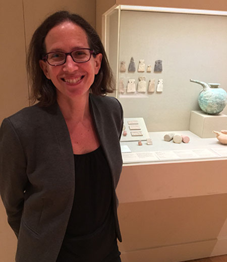 Woman with shoulder-length dark hair, dark glasses, wearing a jacket standing in front of artifacts in a glass.