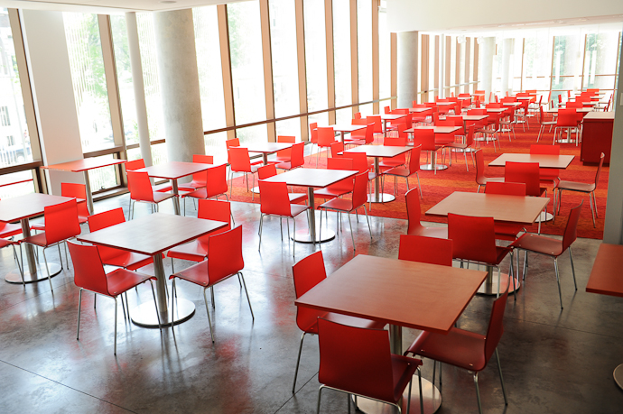 Shows long rectangular room with glass walls and red square tables