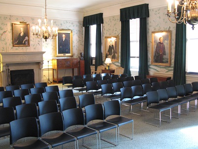 shows elegant room with dark velvet curtains, portraits and ornate wallpaper, a fireplace and rows of black chairs.