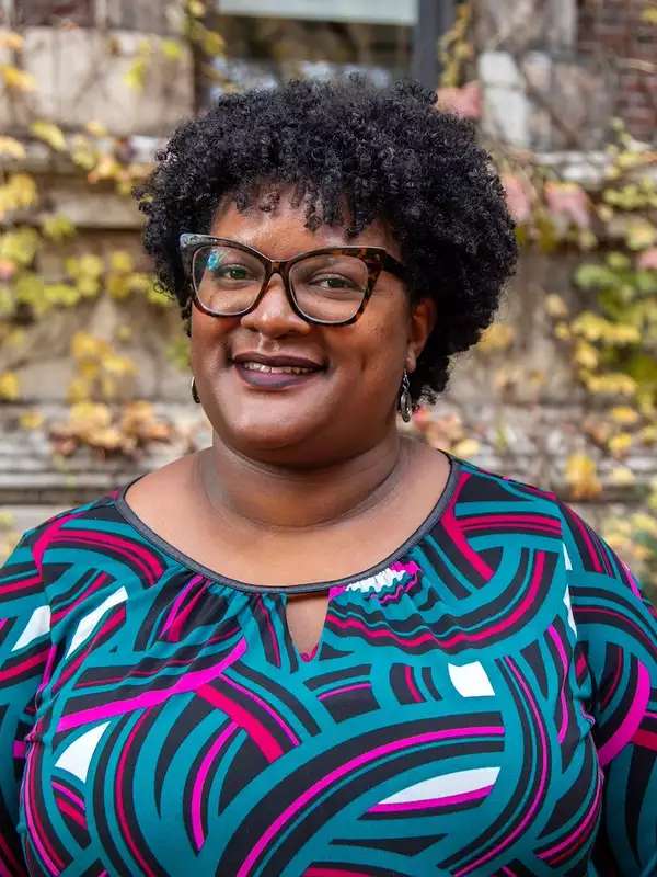 fall background, woman smiling with curly black hair, wearing glasses and a colorful pattered top, smiling