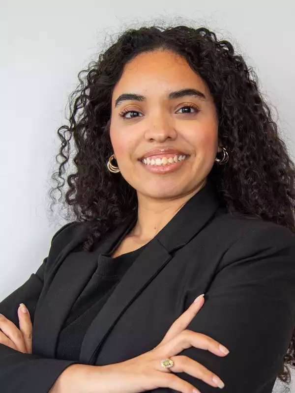 white background, woman with dark brown/black hair, wearing black blazer, smiling, arms crossed