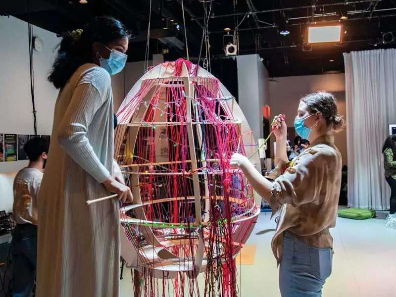 2 students work on a large, egg-shaped object weaving colored yarn