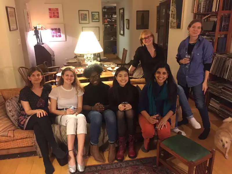 5 young women seated on a couch, 2 older women standing nearby